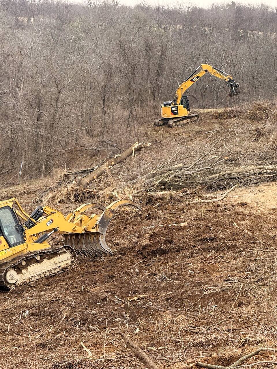 Two tracked machines clearing brush and small timber on a wooded slope along the right-of-way