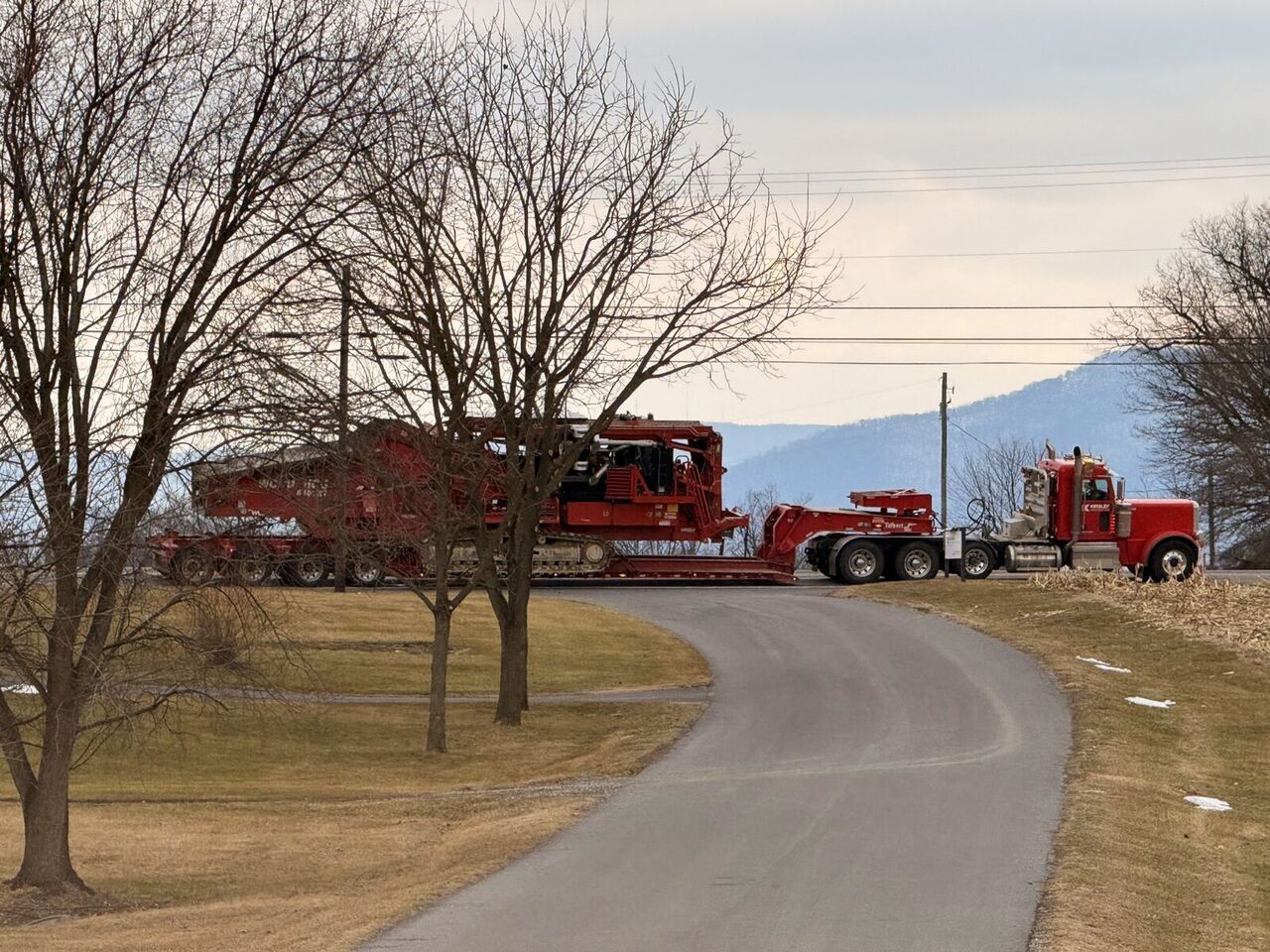 Heavy equipment on a lowboy trailer staged along a rural road with ridgeline in the background