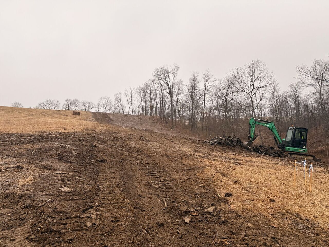 Compact excavator on the graded right-of-way with survey stakes marking the corridor edge